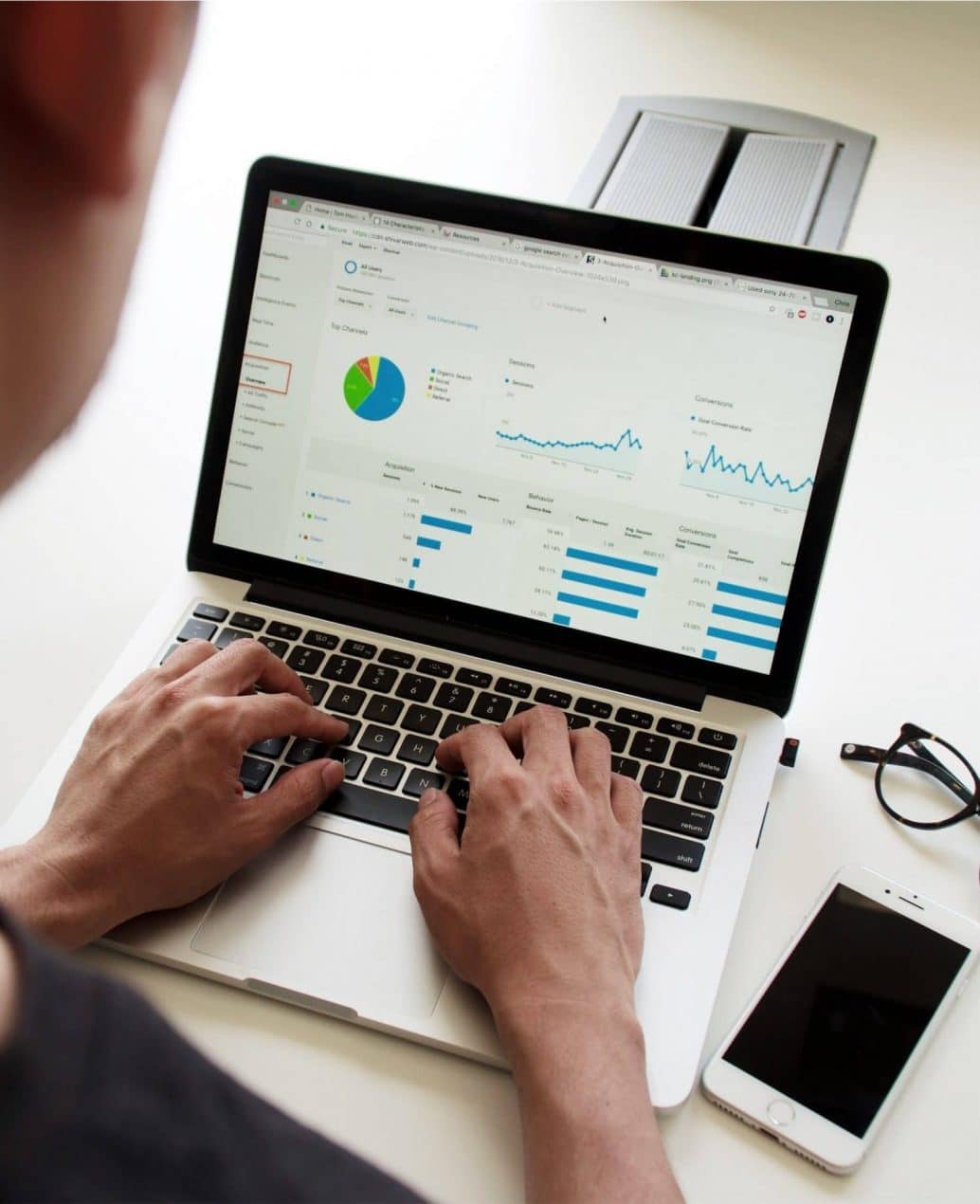 analytics. Das Bild zeigt eine Person, die an einem Laptop arbeitet und Google Analytics-Daten in Form von Diagrammen und Statistiken überprüft, während ein Smartphone und eine Brille auf dem Tisch liegen.