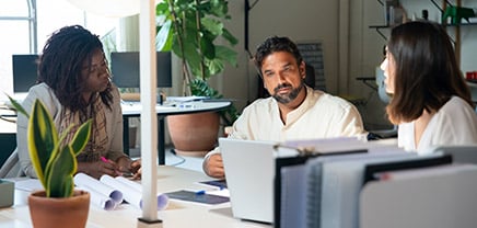 310 310. Das Bild zeigt drei Personen, die in einem Büro an einem Tisch sitzen und sich miteinander unterhalten oder eine Besprechung führen. Es gibt Pflanzen und Schreibtischutensilien im Hintergrund.