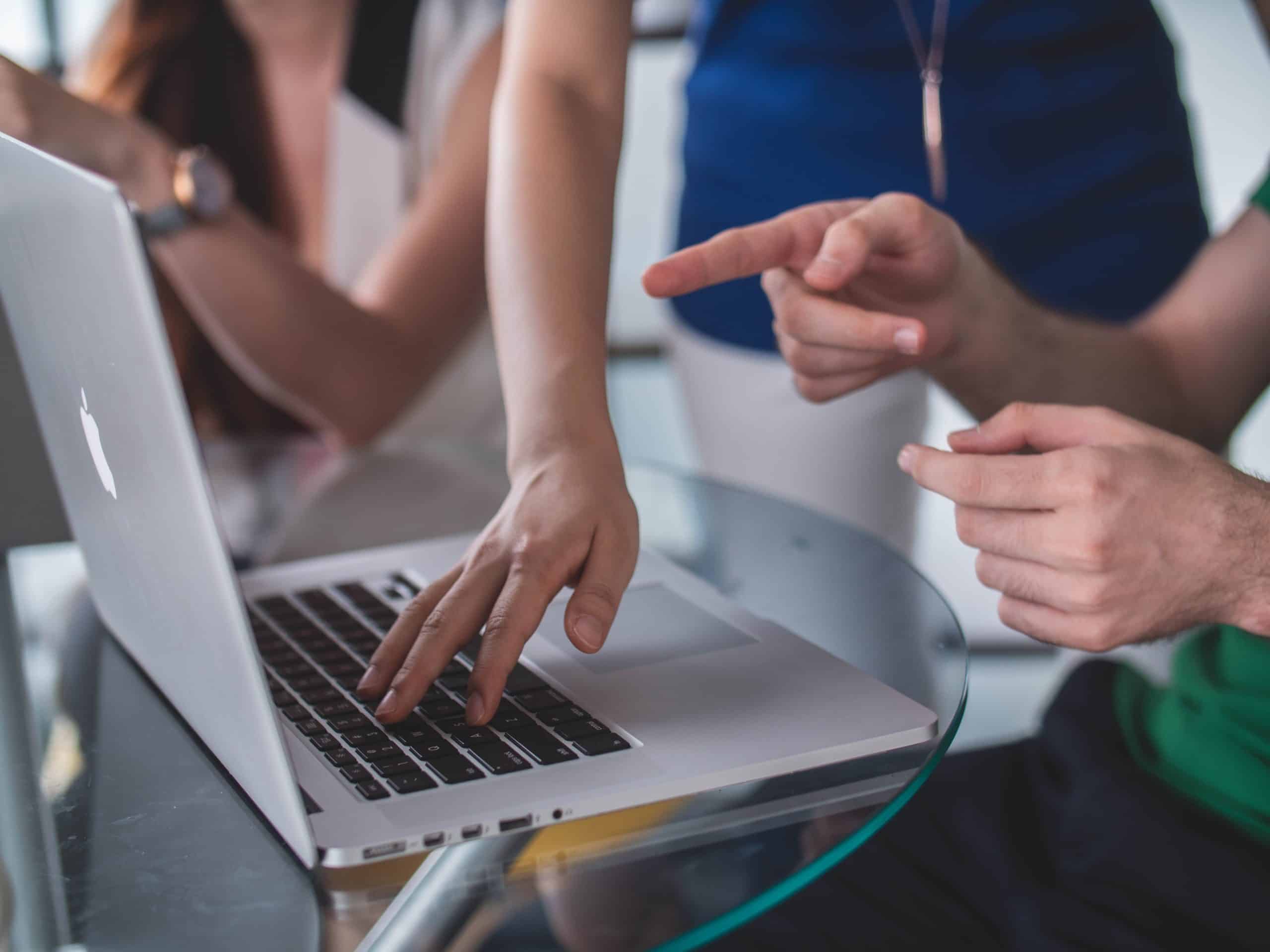 Das Bild zeigt eine Gruppe von Menschen, die zusammen an einem MacBook arbeiten. Eine Person zeigt mit dem Finger auf den Bildschirm, während eine andere Person an der Tastatur tippt.